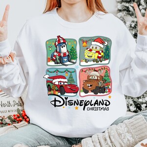 A person is wearing a white sweatshirt with a graphic of four Disney characters from the Cars movie, including a Santa hat, a reindeer, a car, and a snowman, along with the text "Disneyland Christmas".
