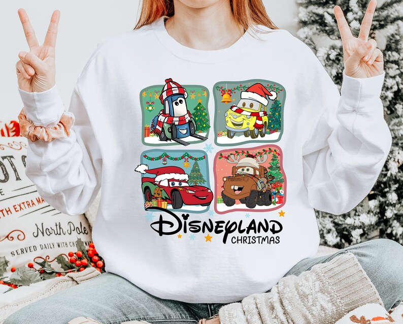 A person is wearing a white sweatshirt with a graphic of four Disney characters from the Cars movie, including a Santa hat, a reindeer, a car, and a snowman, along with the text "Disneyland Christmas".