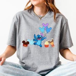 A woman wearing a gray t-shirt with a graphic design featuring a blue cartoon character surrounded by various Disney-themed items such as balloons, Mickey Mouse ears, and a cupcake.
