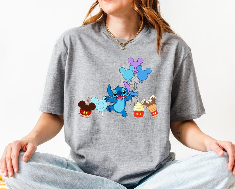 A woman wearing a gray t-shirt with a graphic design featuring a blue cartoon character surrounded by various Disney-themed items such as balloons, Mickey Mouse ears, and a cupcake.