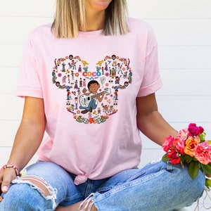 May include: Pink t-shirt with a Mickey Mouse shaped design featuring characters from the Disney Pixar movie Coco. The design includes Miguel playing a guitar and the text "Coco".