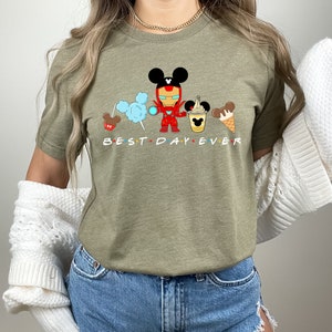 May include: A green t-shirt with a graphic of a cartoon Iron Man character with Mickey Mouse ears, holding a Mickey Mouse ice cream cone. The graphic also includes Mickey Mouse ears with cotton candy, a Mickey Mouse pretzel, and a Mickey Mouse ice cream cone. The text "BEST DAY EVER" is printed below the graphic.
