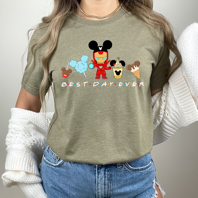 May include: A green t-shirt with a graphic of a cartoon Iron Man character with Mickey Mouse ears, holding a Mickey Mouse ice cream cone. The graphic also includes Mickey Mouse ears with cotton candy, a Mickey Mouse pretzel, and a Mickey Mouse ice cream cone. The text "BEST DAY EVER" is printed below the graphic.