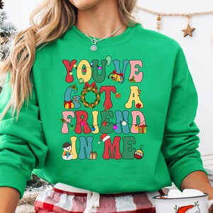 A woman wearing a green sweatshirt with a colorful text design on it, standing in front of a Christmas tree.