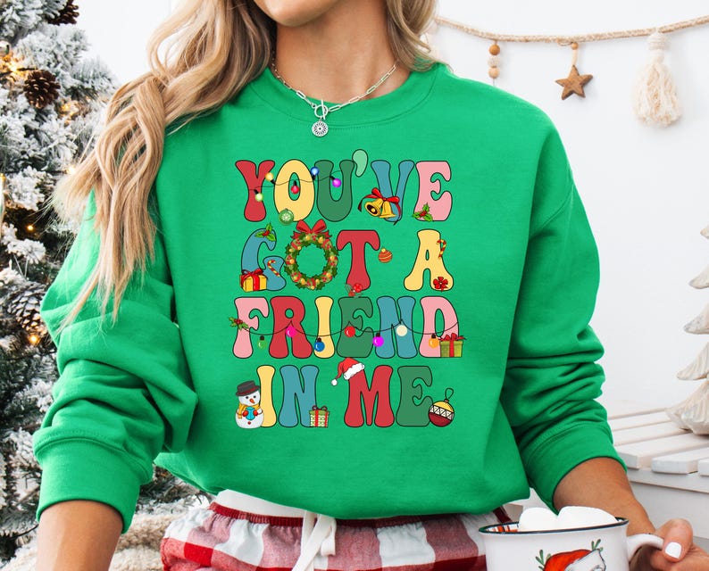A woman wearing a green sweatshirt with a colorful text design on it, standing in front of a Christmas tree.