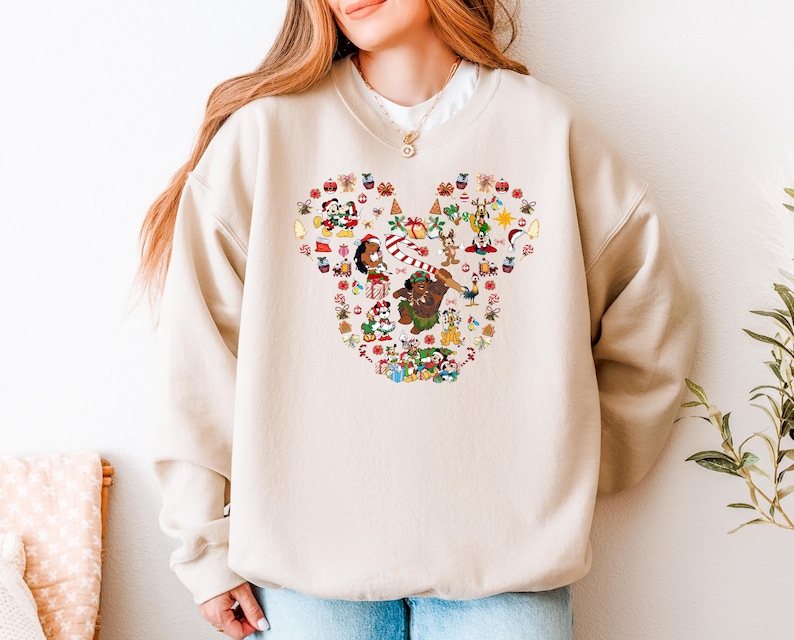 A woman wearing a beige sweater with a large, colorful, cartoonish design of various characters and objects, including a snowman, reindeer, and other festive elements.