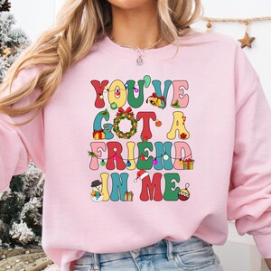 A woman wearing a pink sweatshirt with a colorful graphic design that says "You've Got a FRIEND in Me" in a playful, festive style.