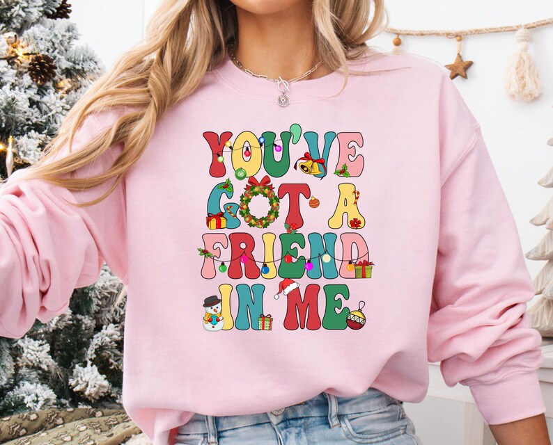 A woman wearing a pink sweatshirt with a colorful graphic design that says "You've Got a FRIEND in Me" in a playful, festive style.