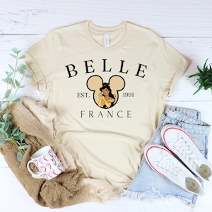 Puede incluir: Una camiseta beige con un dise&ntilde;o gr&aacute;fico negro que presenta a Bella de La Bella y la Bestia dentro de una silueta de oreja de Mickey Mouse. El texto "BELLE EST. 1991 FRANCE" est&aacute; impreso debajo del gr&aacute;fico.