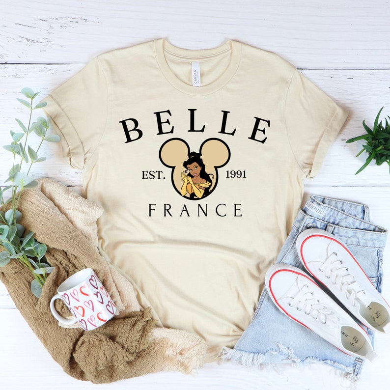 Puede incluir: Una camiseta beige con un dise&ntilde;o gr&aacute;fico negro que presenta a Bella de La Bella y la Bestia dentro de una silueta de oreja de Mickey Mouse. El texto "BELLE EST. 1991 FRANCE" est&aacute; impreso debajo del gr&aacute;fico.