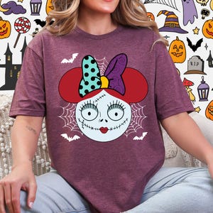 A woman wearing a purple t-shirt with a cartoon character design featuring a bow on her head and a spooky Halloween-themed background.