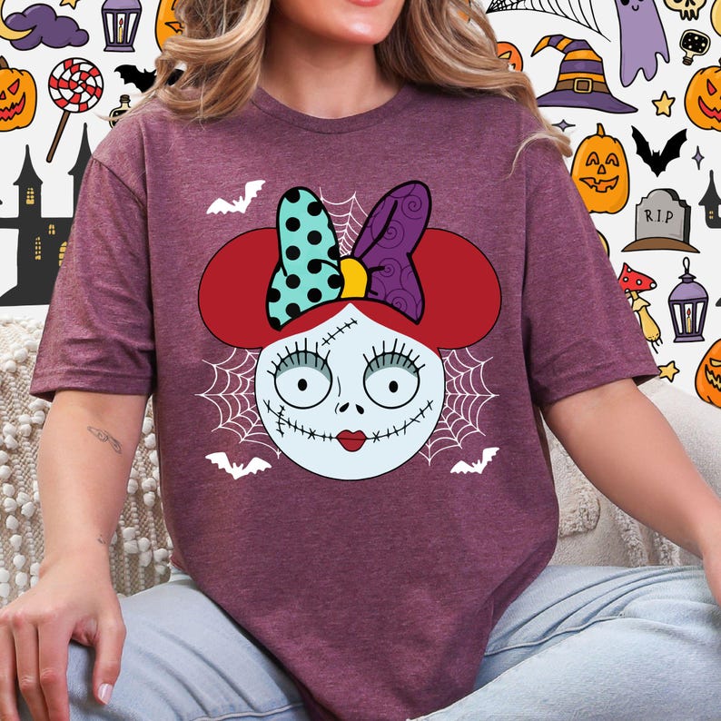 A woman wearing a purple t-shirt with a cartoon character design featuring a bow on her head and a spooky Halloween-themed background.