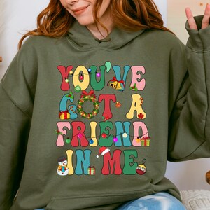 A woman wearing a green hoodie with a colorful graphic design that says "YOU'VE GOT A FRIEND IN ME" and includes various Christmas-themed elements such as a snowman, presents, and a Christmas tree.