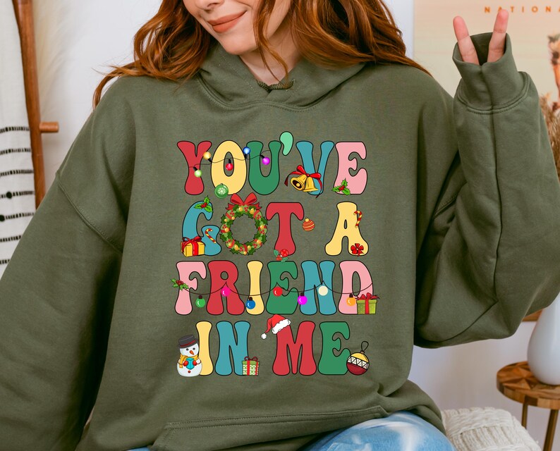 A woman wearing a green hoodie with a colorful graphic design that says "YOU'VE GOT A FRIEND IN ME" and includes various Christmas-themed elements such as a snowman, presents, and a Christmas tree.
