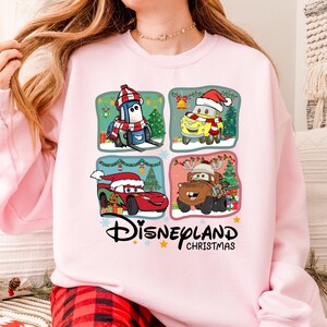 A woman wearing a pink sweatshirt with a collage of four cartoon car characters from the Disney movie Cars, including a Santa Claus car, a reindeer car, a car with a Christmas tree, and a car with a snowman.