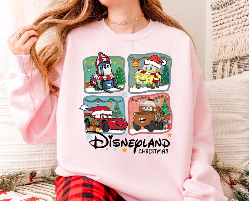A woman wearing a pink sweatshirt with a collage of four cartoon car characters from the Disney movie Cars, including a Santa Claus car, a reindeer car, a car with a Christmas tree, and a car with a snowman.