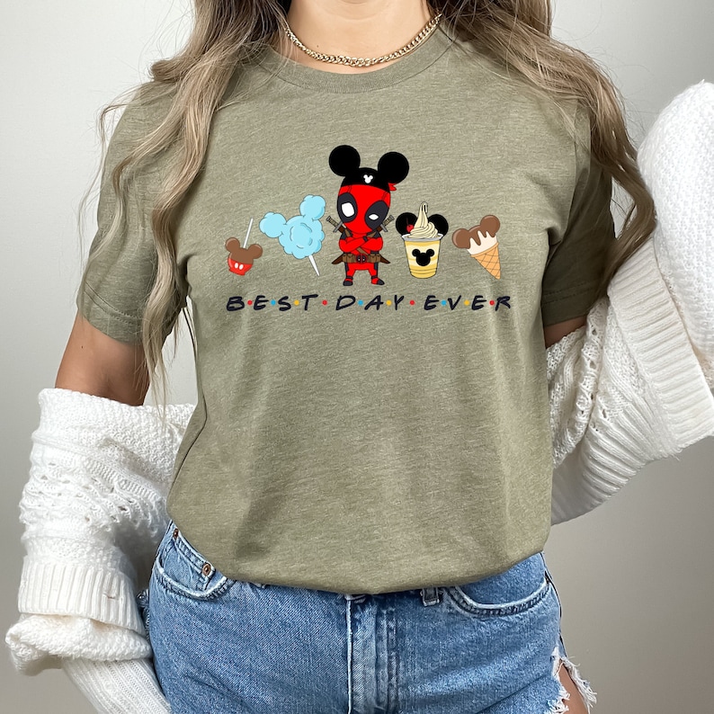 May include: A green t-shirt with a cartoon image of Deadpool wearing Mickey Mouse ears and holding a sword. The shirt has the text "BEST DAY EVER" printed on it.
