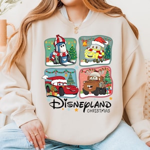 A woman wearing a white sweatshirt with a graphic design featuring four cartoon cars from the Disney movie Cars, along with Christmas-themed elements.