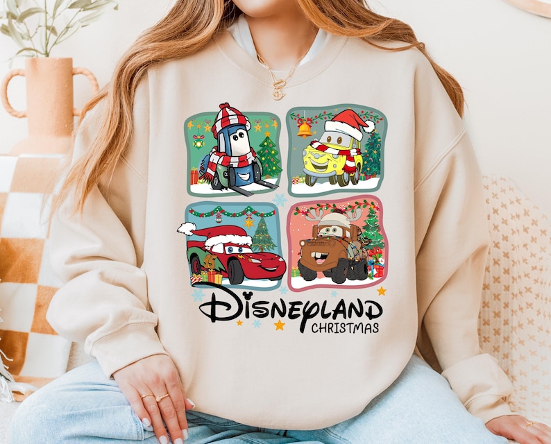 A woman wearing a white sweatshirt with a graphic design featuring four cartoon cars from the Disney movie Cars, along with Christmas-themed elements.