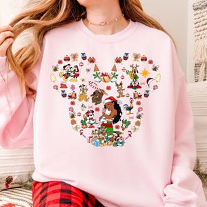 A woman wearing a pink sweatshirt with a colorful design featuring various Disney characters and holiday-themed elements.