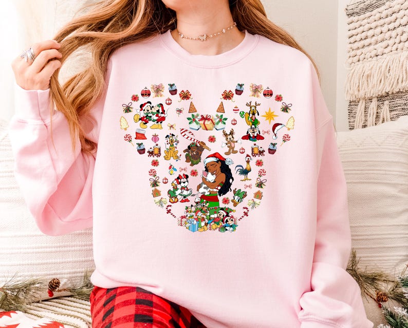 A woman wearing a pink sweatshirt with a colorful design featuring various Disney characters and holiday-themed elements.