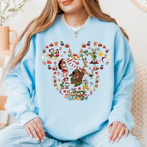 A woman wearing a light blue sweatshirt with a colorful design featuring various Disney characters and holiday-themed elements.