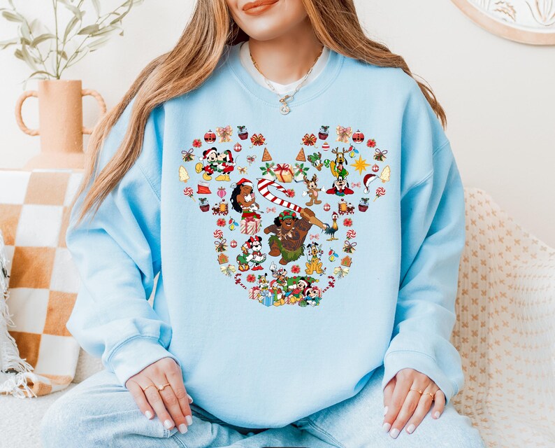 A woman wearing a light blue sweatshirt with a colorful design featuring various Disney characters and holiday-themed elements.