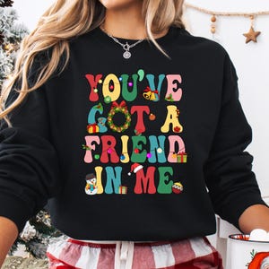 A woman wearing a black sweatshirt with a colorful graphic design that says "YOU'VE GOT A FRIEND IN ME" in a playful, festive style.
