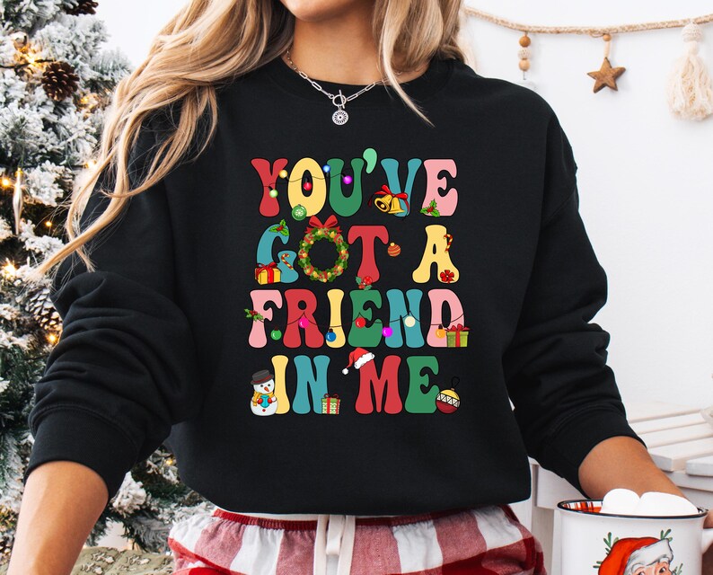 A woman wearing a black sweatshirt with a colorful graphic design that says "YOU'VE GOT A FRIEND IN ME" in a playful, festive style.