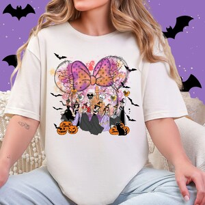 May include: Cream-colored t-shirt featuring a Halloween-themed graphic. The design includes a large purple bow with orange polka dots, a castle, bats, pumpkins, and cartoon characters in costumes. The shirt is a casual, festive apparel item.