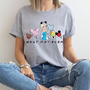 Puede incluir: Camiseta gris con un gr&aacute;fico de una princesa Disney de dibujos animados sosteniendo un algod&oacute;n de az&uacute;car azul, un cono de helado amarillo y un helado de Mickey Mouse rojo. El texto "BEST DAY EVER" est&aacute; impreso debajo del gr&aacute;fico.
