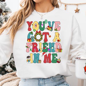 A woman wearing a white sweatshirt with a colorful text design that says "You've Got a Friend in Me" and a Santa Claus mug.