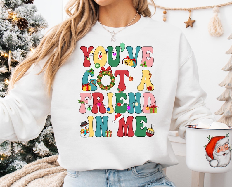 A woman wearing a white sweatshirt with a colorful text design that says "You've Got a Friend in Me" and a Santa Claus mug.