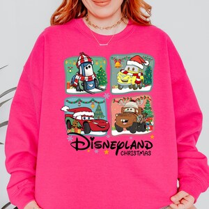 A woman wearing a pink sweatshirt with a collage of four Disney Cars characters from the movie "Disneyland Christmas" on it.