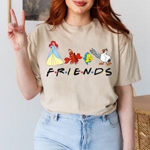 K&ouml;nnte beinhalten: Ein beiges T-Shirt mit einem grafischen Aufdruck von Figuren aus Arielle, die Meerjungfrau, darunter Arielle, Sebastian, Flounder und Scuttle. Der Text "FRIENDS" ist unter den Figuren aufgedruckt.