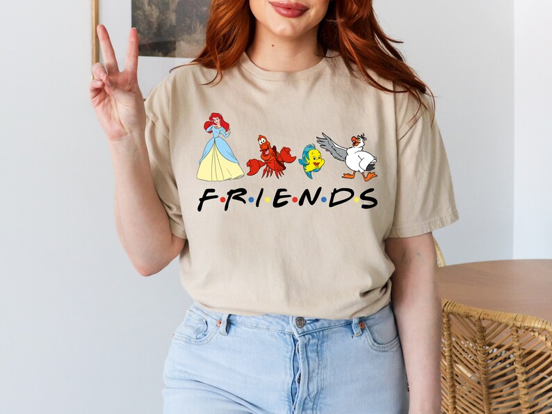 K&ouml;nnte beinhalten: Ein beiges T-Shirt mit einem grafischen Aufdruck von Figuren aus Arielle, die Meerjungfrau, darunter Arielle, Sebastian, Flounder und Scuttle. Der Text "FRIENDS" ist unter den Figuren aufgedruckt.