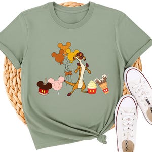 May include: Sage green t-shirt featuring a cartoon meerkat holding balloons, with Mickey Mouse-shaped balloons, cotton candy, ice cream, and a cupcake. White canvas sneakers are next to the shirt. The shirt is knotted at the bottom.