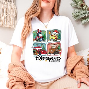 A woman wearing a white t-shirt with a Disney-themed graphic of four cartoon cars, including a Santa Claus car, is sitting in a room decorated for Christmas.