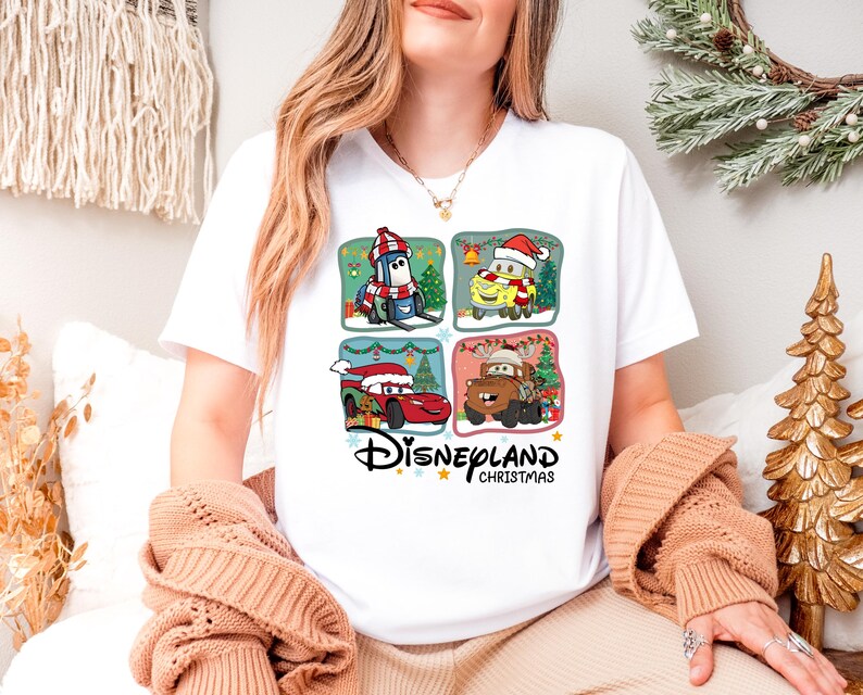 A woman wearing a white t-shirt with a Disney-themed graphic of four cartoon cars, including a Santa Claus car, is sitting in a room decorated for Christmas.