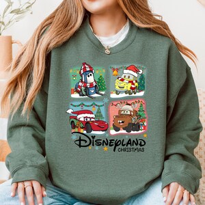 A woman wearing a green sweatshirt with a collage of four Disney Cars characters from the Cars movie, including a Santa Claus car, a reindeer car, a car with a Christmas tree, and a car with a Christmas tree.