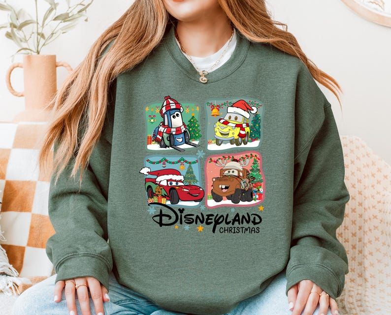 A woman wearing a green sweatshirt with a collage of four Disney Cars characters from the Cars movie, including a Santa Claus car, a reindeer car, a car with a Christmas tree, and a car with a Christmas tree.