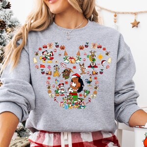 A woman wearing a gray sweatshirt with a heart-shaped design featuring various Disney characters and holiday-themed elements.