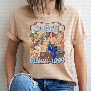May include: A tan t-shirt with a vintage-style graphic of characters from the Disney movie Tarzan. The graphic includes the text "Disney Tarzan Since 1999".
