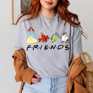 K&ouml;nnte beinhalten: Ein graues T-Shirt mit einem Aufdruck von Figuren aus Arielle, die Meerjungfrau, darunter Arielle, Sebastian, Flounder und Scuttle. Der Text "FRIENDS" befindet sich unter den Figuren, wobei jeder Buchstabe durch einen farbigen Punkt getrennt ist.