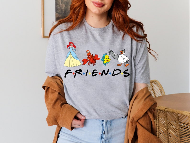 K&ouml;nnte beinhalten: Ein graues T-Shirt mit einem Aufdruck von Figuren aus Arielle, die Meerjungfrau, darunter Arielle, Sebastian, Flounder und Scuttle. Der Text "FRIENDS" befindet sich unter den Figuren, wobei jeder Buchstabe durch einen farbigen Punkt getrennt ist.