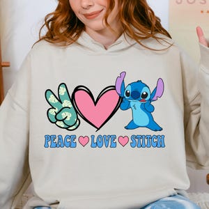 A woman wearing a white hoodie with a graphic design featuring a character from the Lilo & Stitch movie franchise, along with the text "PEACE LOVE STITCH".