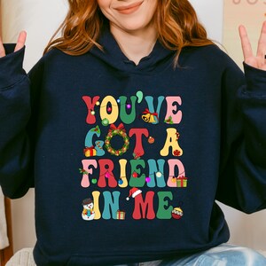 A woman wearing a navy blue hoodie with a colorful graphic design that says "You've Got a Friend in Me" and includes various Christmas-themed elements such as a snowman, presents, and a Christmas tree.