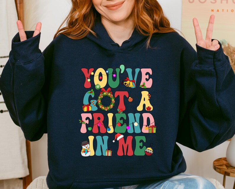 A woman wearing a navy blue hoodie with a colorful graphic design that says "You've Got a Friend in Me" and includes various Christmas-themed elements such as a snowman, presents, and a Christmas tree.