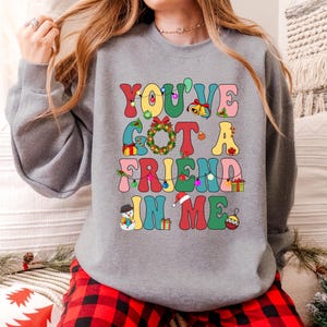 A woman wearing a gray sweatshirt with a colorful text design that says "YOU'VE GOT A FRIEND IN ME" printed on it.