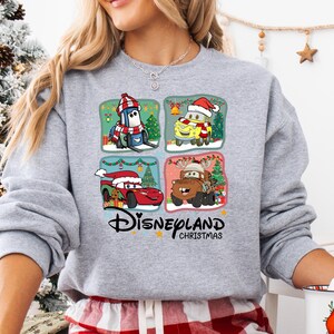 A woman wearing a gray sweatshirt with a Disneyland Christmas-themed design featuring four cartoon cars and a Christmas tree.
